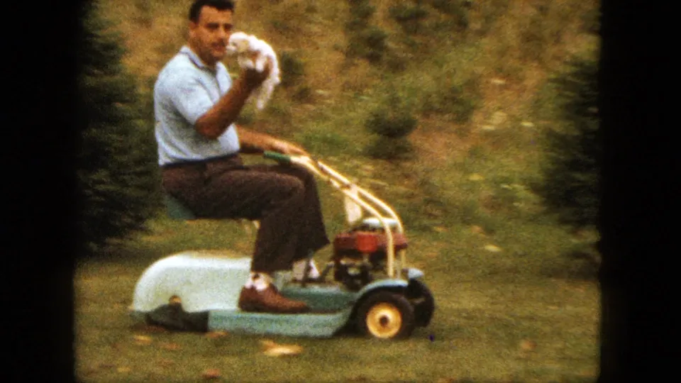 The man turns toward the camera on the lawnmower and raises the puppy near his face in the 1963 home-movie frame.