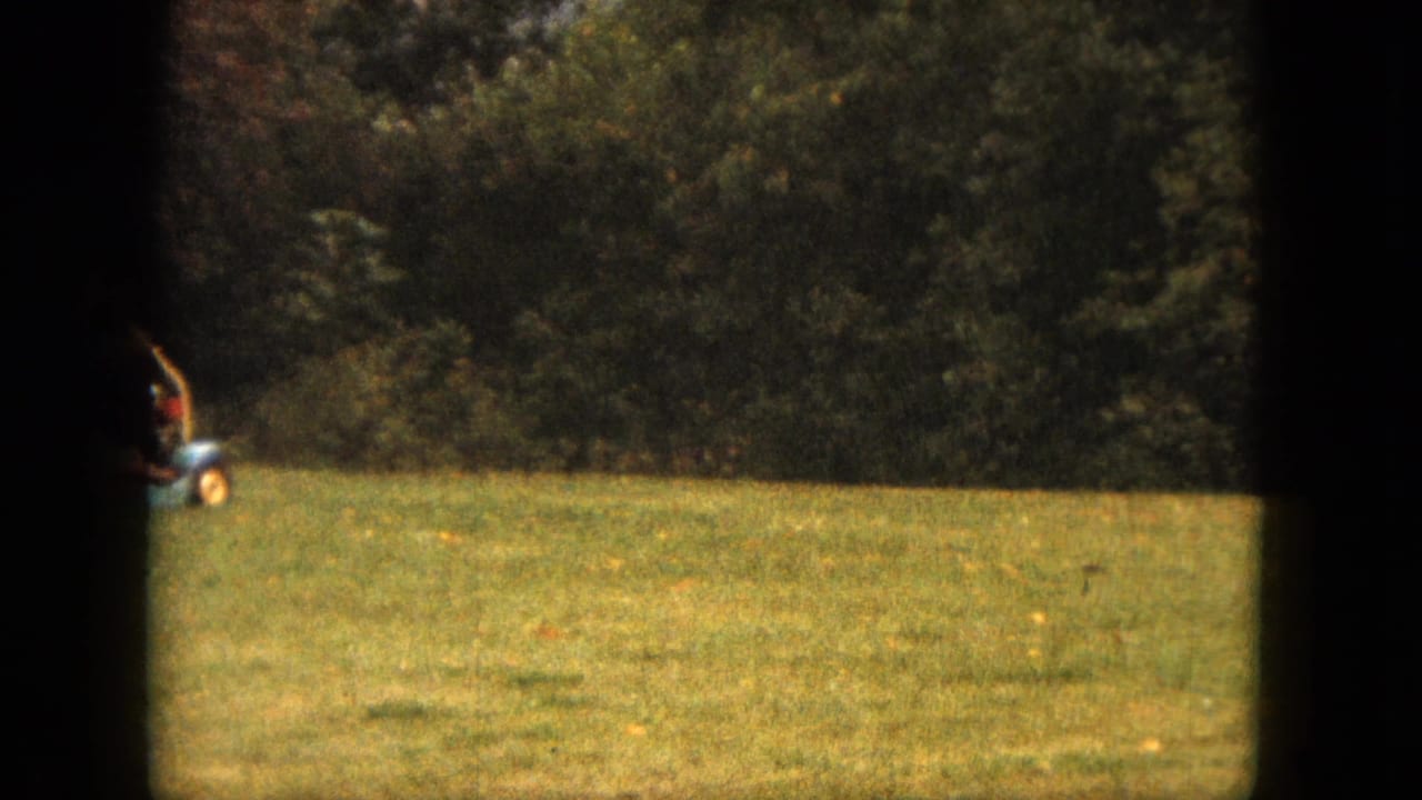 Man on a small riding lawn mower crossing a green backyard near trees.
