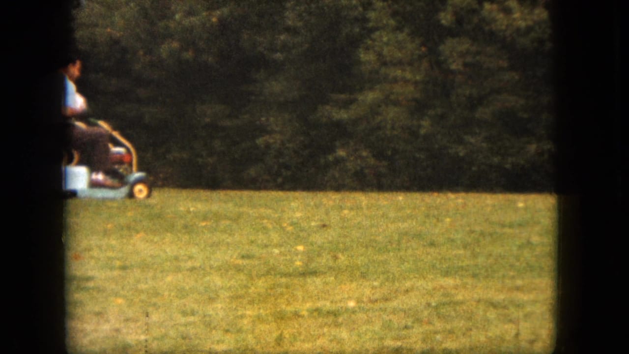 A distant 1963 Saint Paul backyard view shows a man on a small riding mower against a tree-lined lawn.