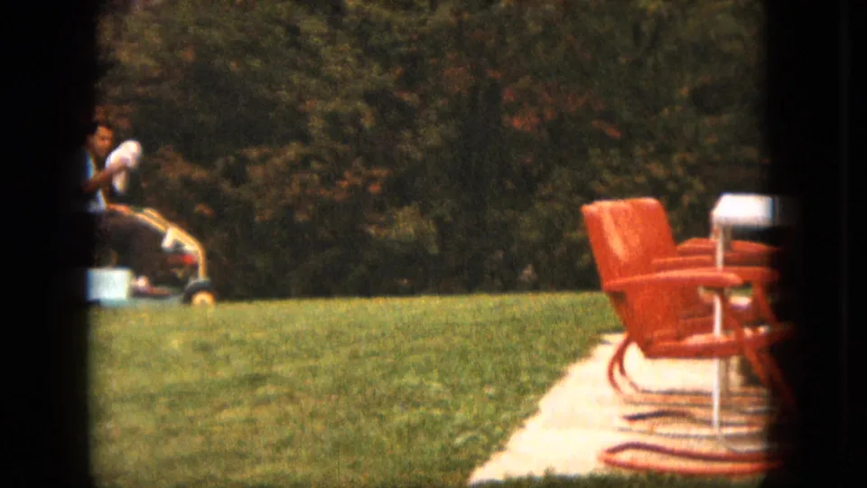 A man rides a small mower across a Saint Paul backyard near red patio chairs, a garden hose, and a paved patio area.