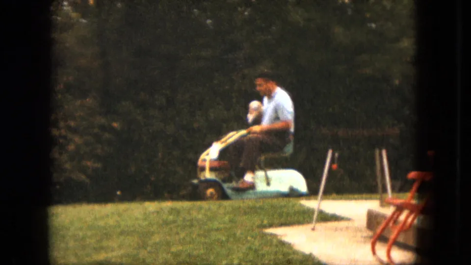 A man in a light blue shirt rides a small mower across a Saint Paul backyard beside a paved patio and red lawn chair.