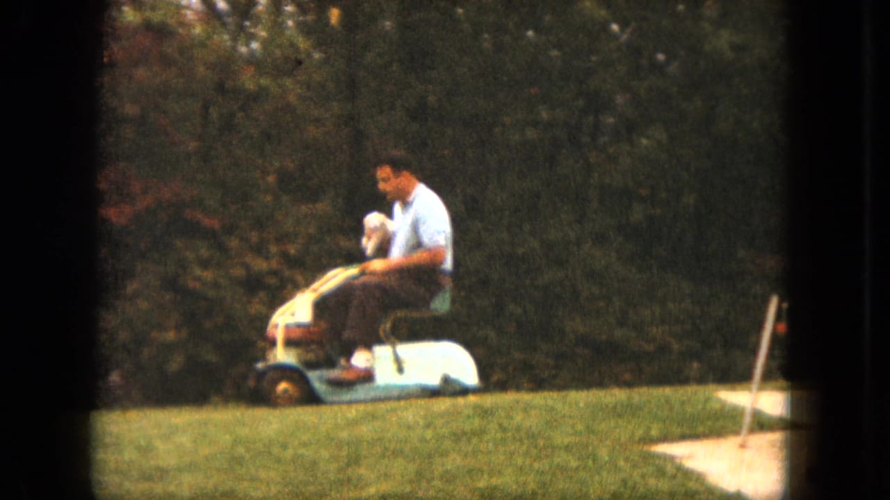 Man rides a small lawn mower across grass while holding a white dog.
