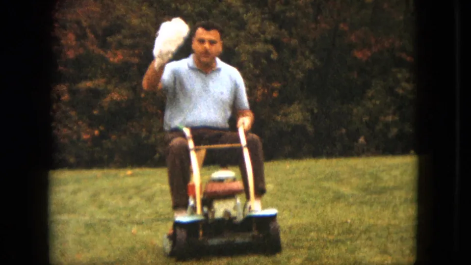 The man rides a vintage mower in a Saint Paul yard and lifts the white puppy toward the camera.