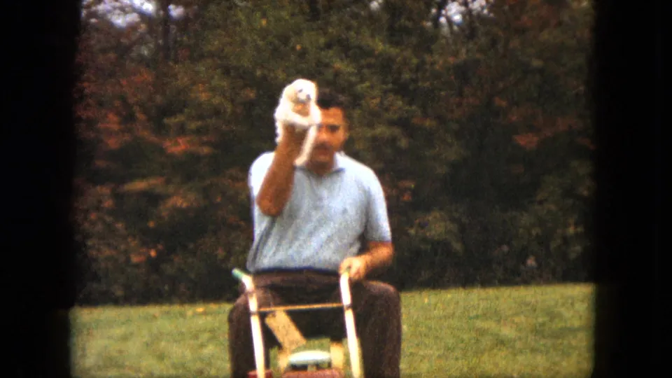 A closer 8mm frame shows the man seated on the mower, holding the small white puppy forward in a grassy Saint Paul setting.