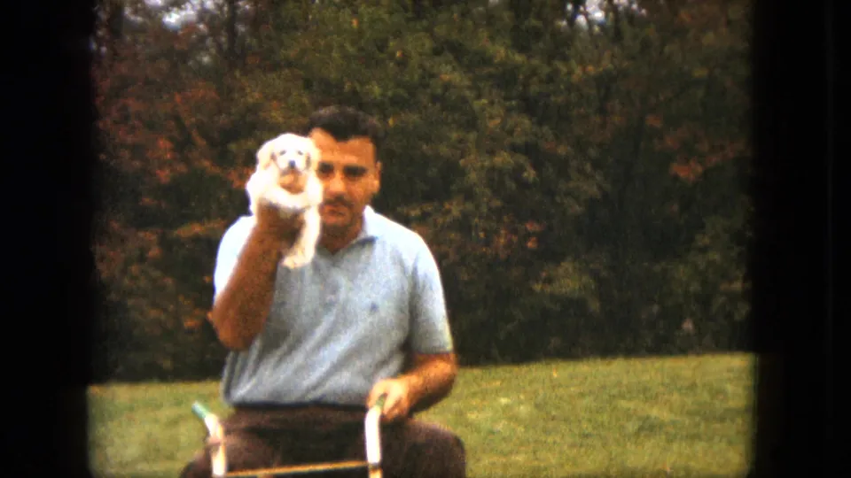 The man looks toward the camera from the ride-on mower, holding the white puppy beside his head against trees and grass.