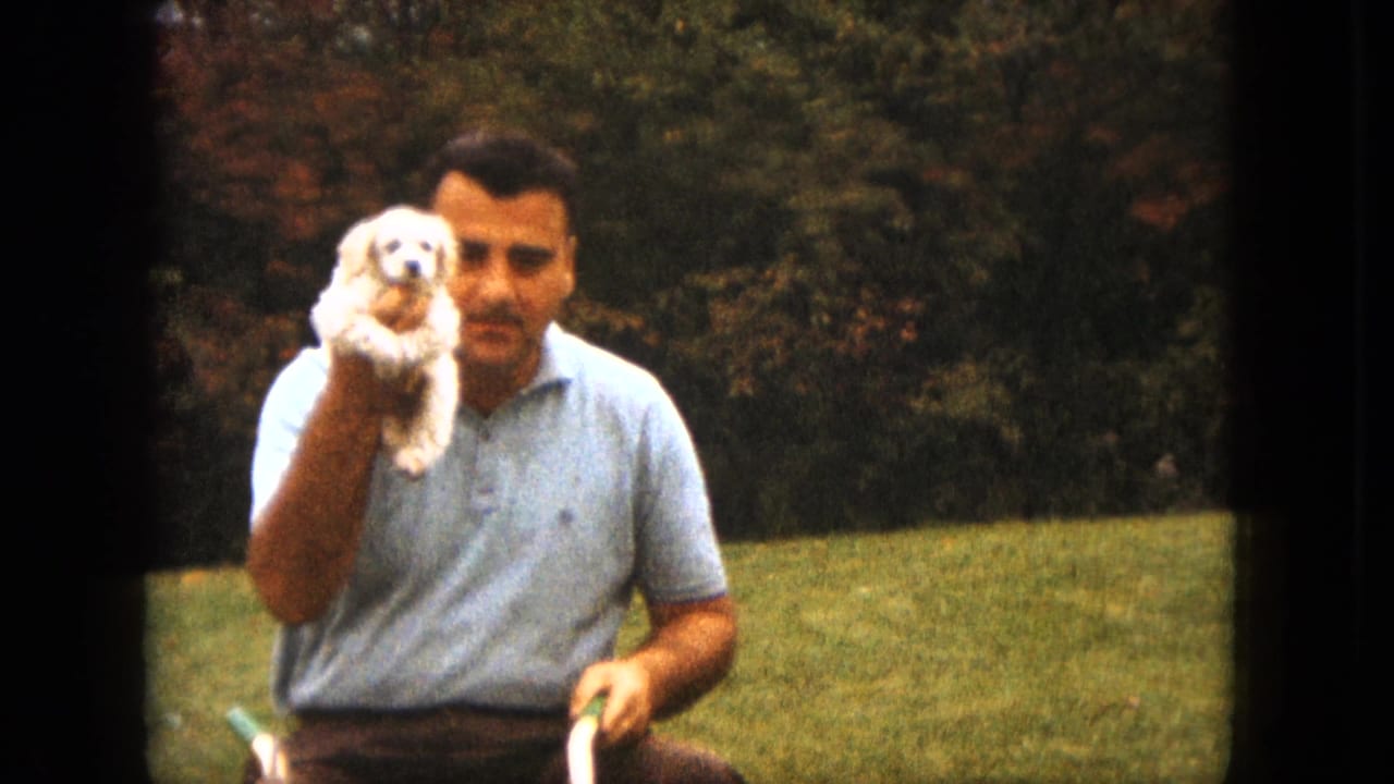 Man in a light blue shirt holds a small white puppy in a grassy yard.