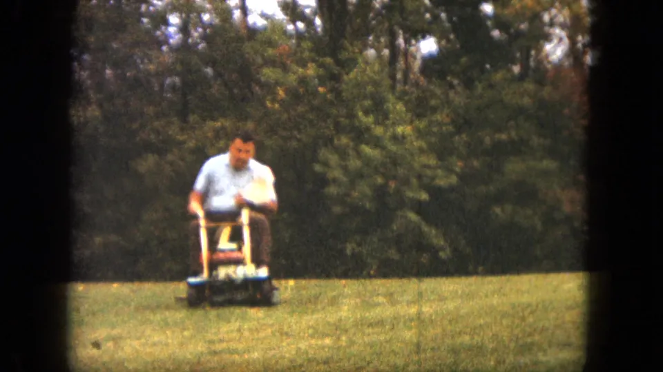 The man rides a small lawnmower across the Saint Paul yard, with open grass and trees in the background.