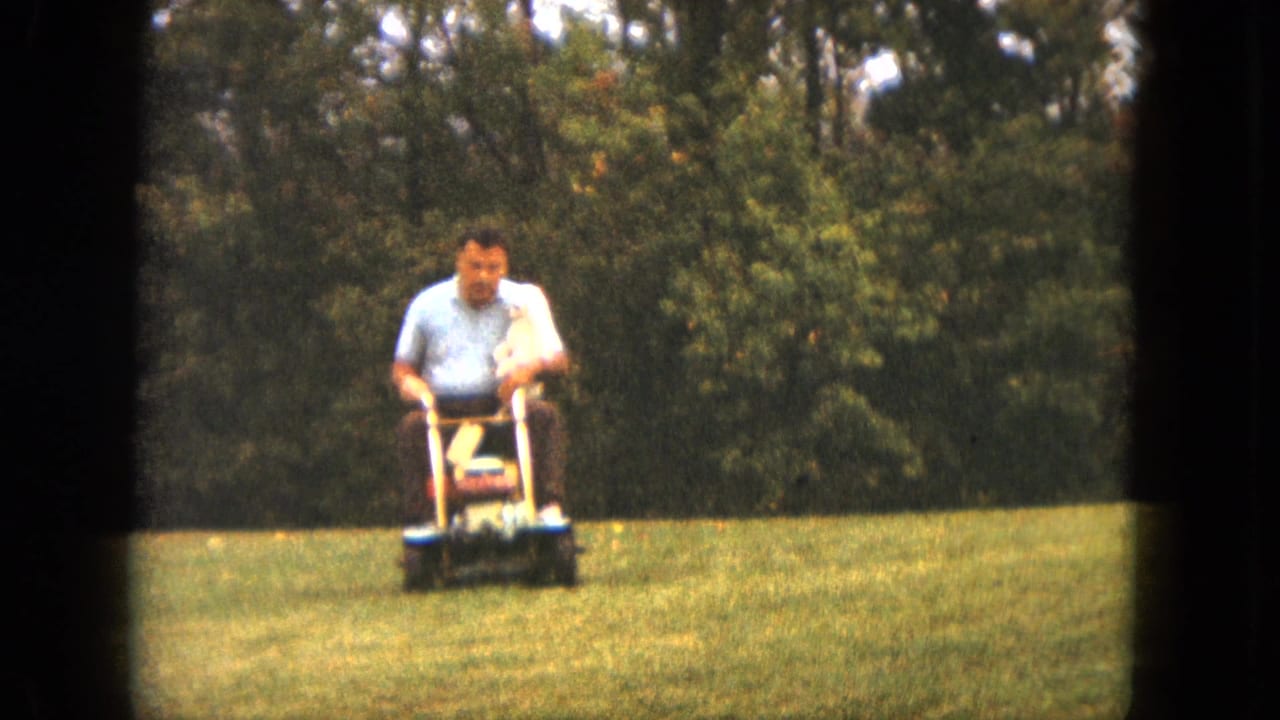 Man rides a mower across a green lawn with a small white dog in his lap.