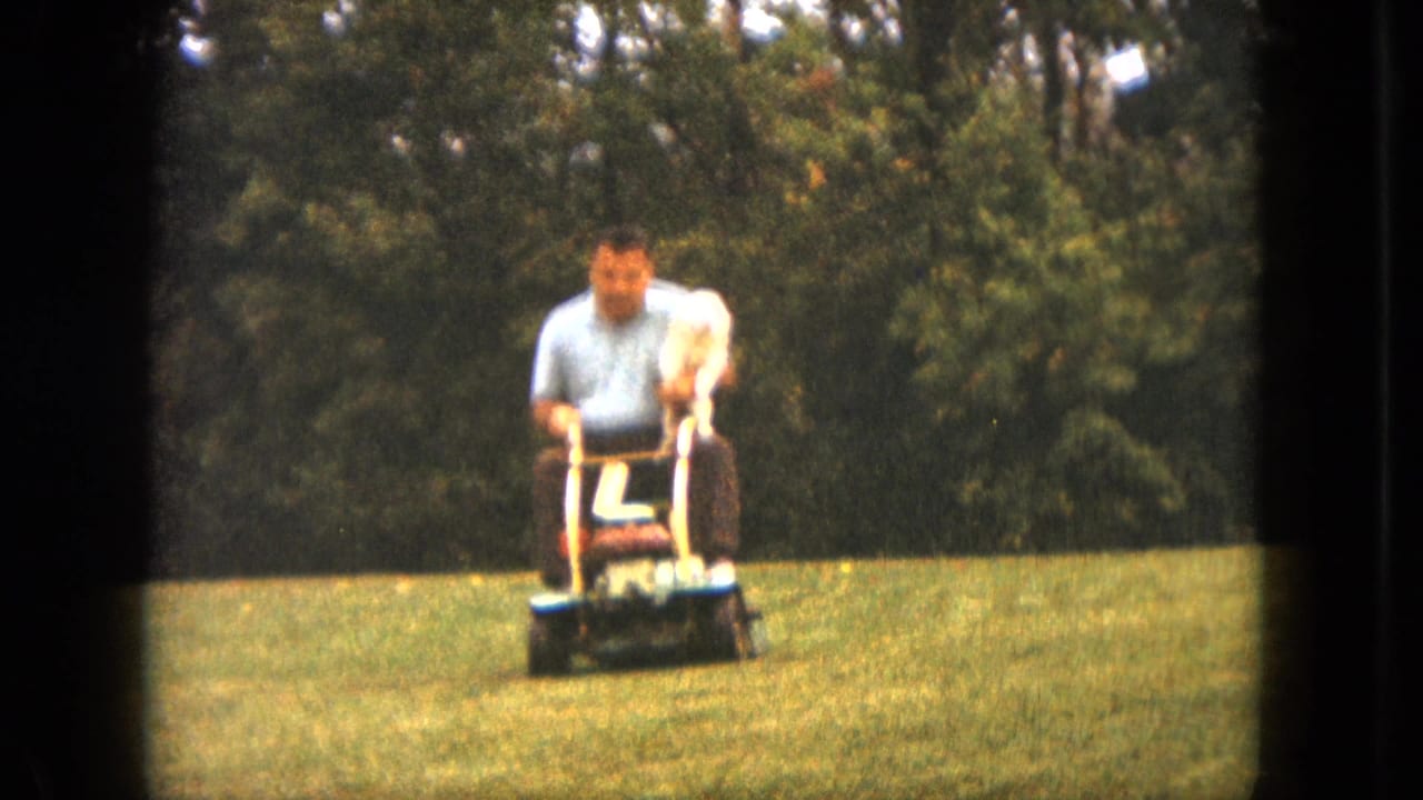 The man continues across the Saint Paul lawn on a ride-on mower, with the small white dog perched calmly in front of him.