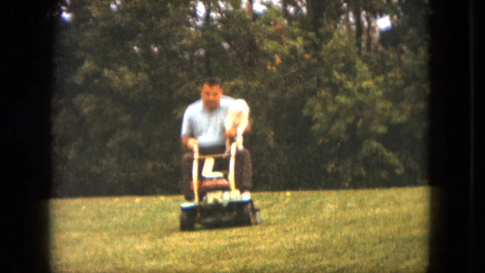 The man continues across the Saint Paul lawn on a ride-on mower, with the small white dog perched calmly in front of him.