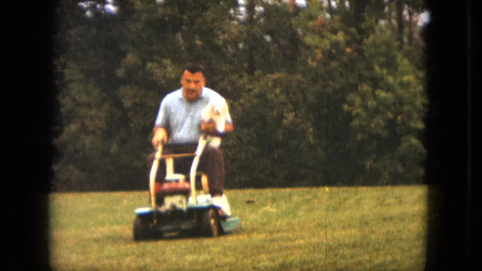A closer 8mm view shows the man and dog on the ride-on mower, framed by green grass and a dense tree backdrop in Saint Paul.