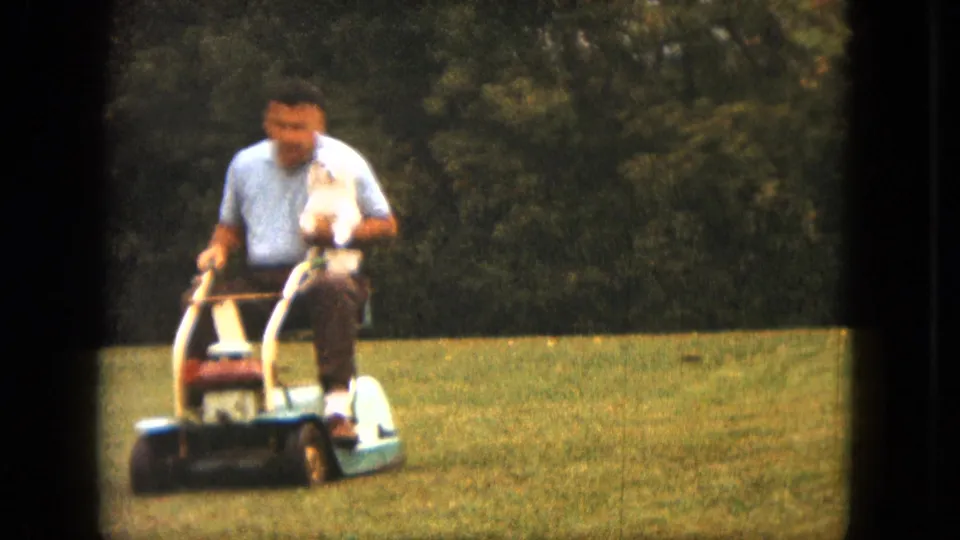 Near the end of the clip, the man rides nearer to the camera on the mower while holding the small white dog in his lap.