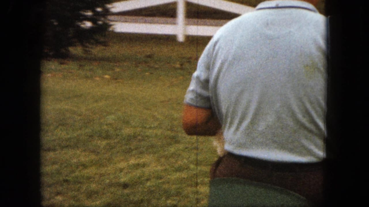 Man seen from behind riding across grass near a white rail fence.