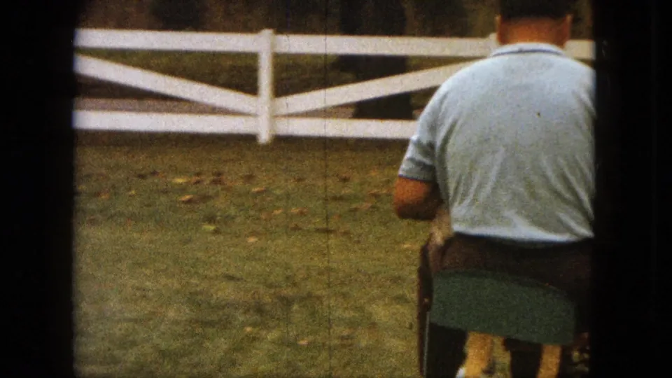 1963 Saint Paul home-movie view of a man riding a small mower across a lawn in front of a white fence.