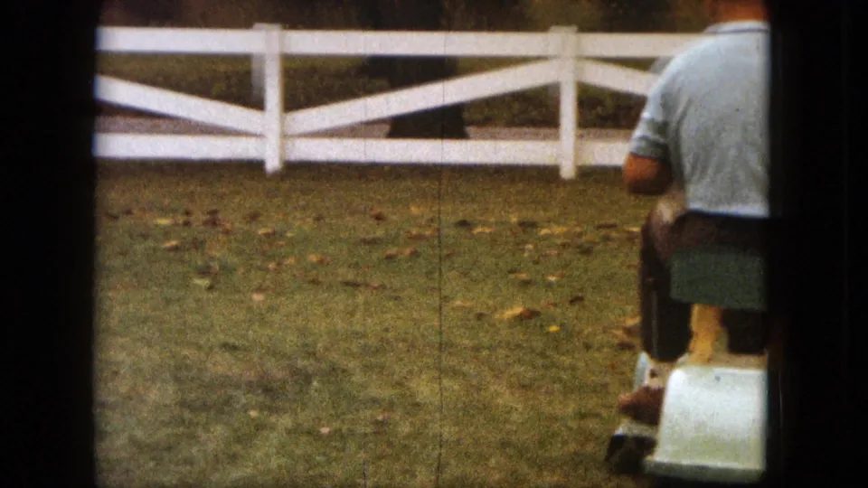 1963 Saint Paul 8mm frame with the rider partly cropped as the mower passes along the grassy yard near a white fence.