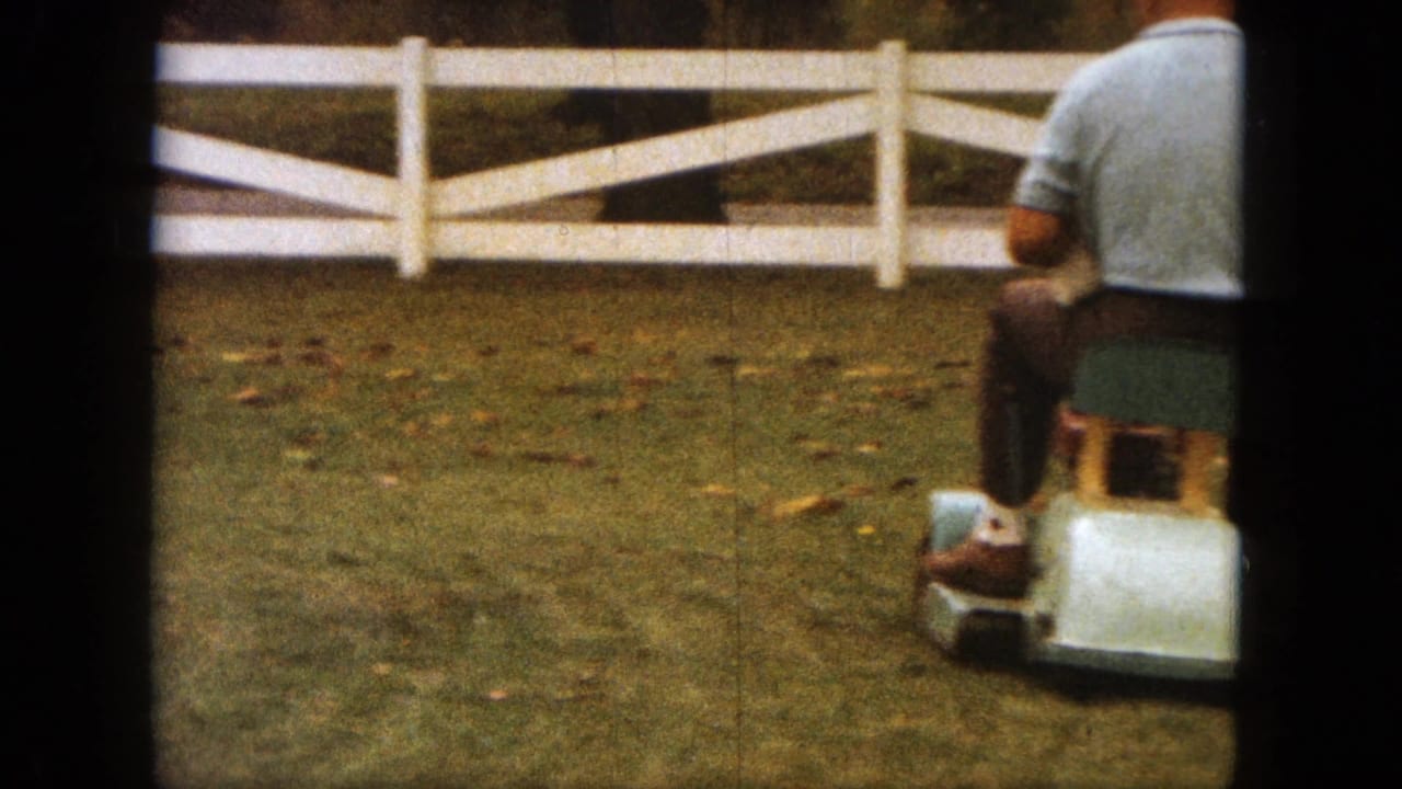 In this 1963 Saint Paul home-movie frame, a man on a small ride-on mower moves across a grassy yard beside a white rail fence.