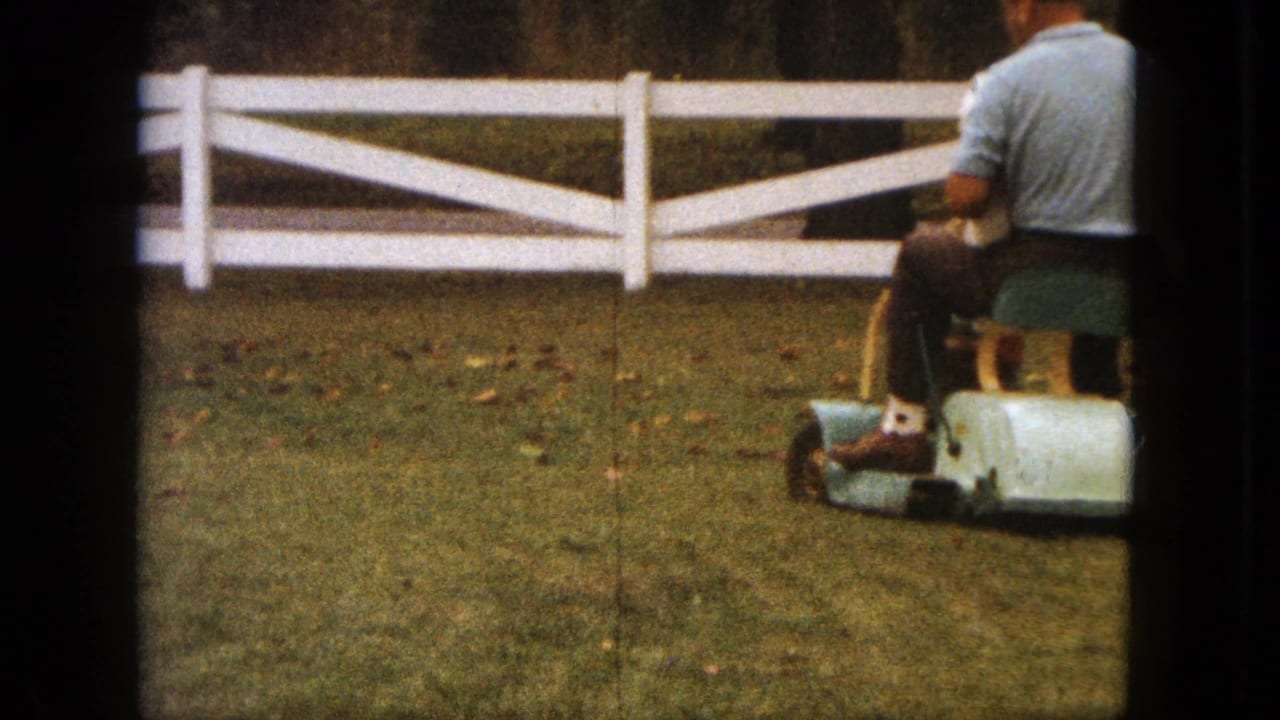The man continues across the Saint Paul yard on a small ride-on mower, with the white fence and lawn filling the background.