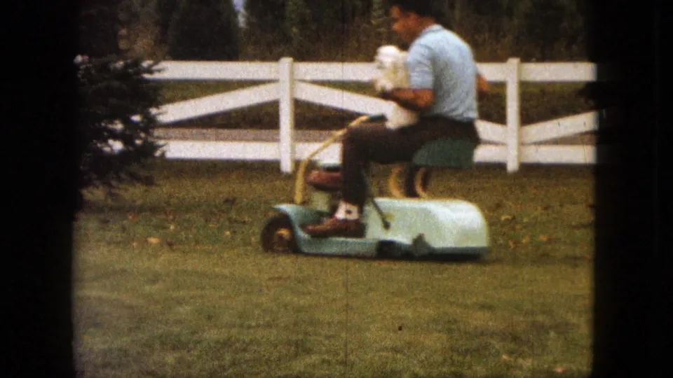 A man in a light blue shirt rides a small mower while carrying a small white dog in his lap in the 1963 Saint Paul yard.