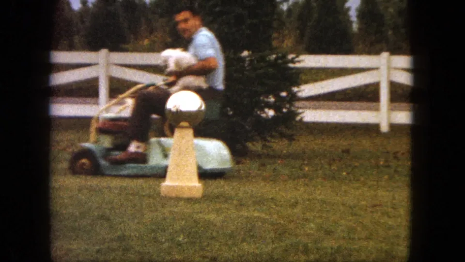 The man rides past a shrub and gold-colored yard ornament while holding the small white dog on the mower in Saint Paul, 1963.
