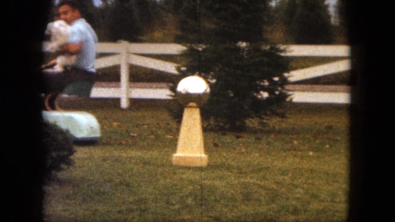 In 1963 Saint Paul, a man in a light blue shirt holds a small white dog while riding a mower across a grassy suburban yard.