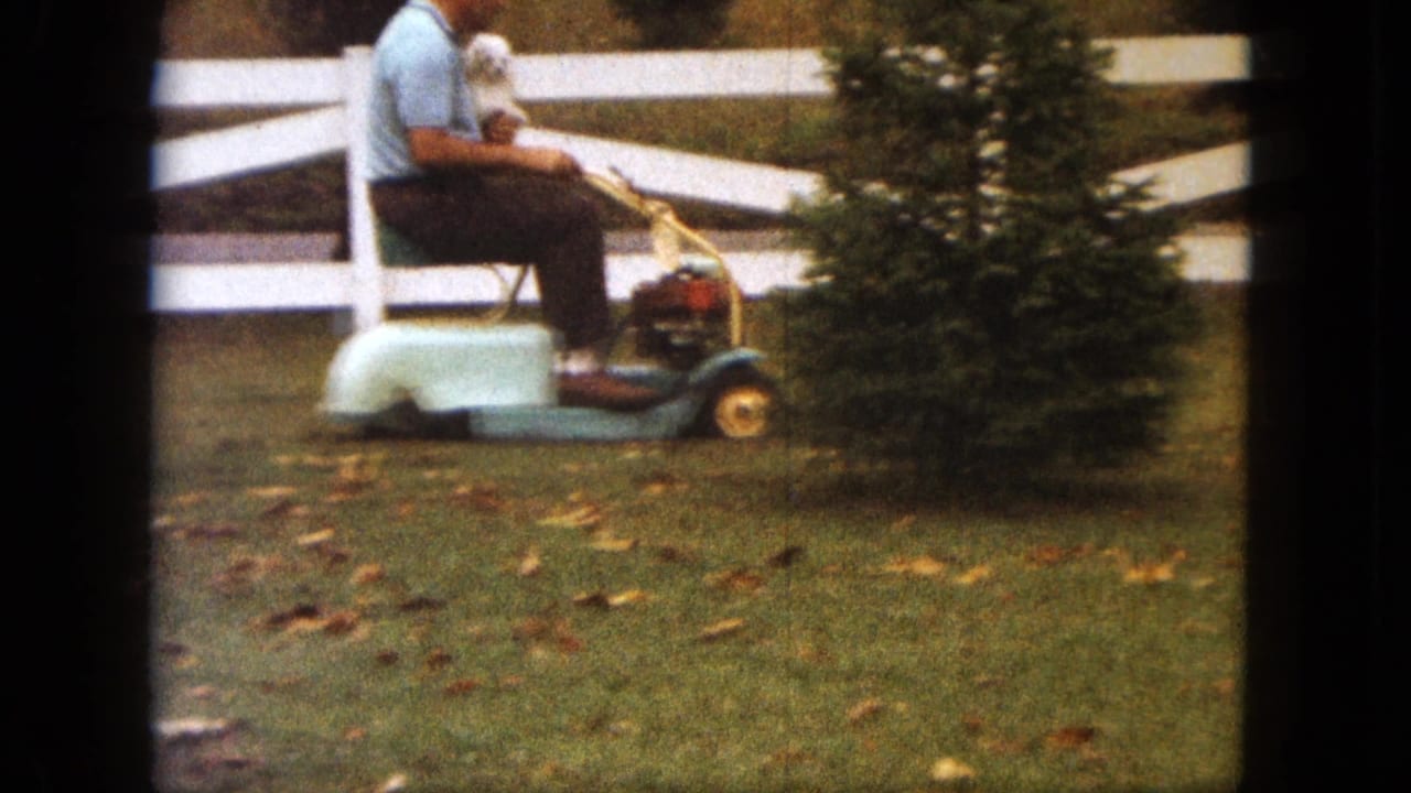 Side view from the 1963 Saint Paul home movie shows the man holding the small dog while riding a compact lawn mower past a white fence.