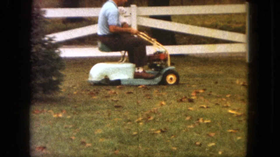 The Saint Paul yard scene continues as the man rides the mower across grass scattered with autumn leaves, with the small dog partly visible in his lap.
