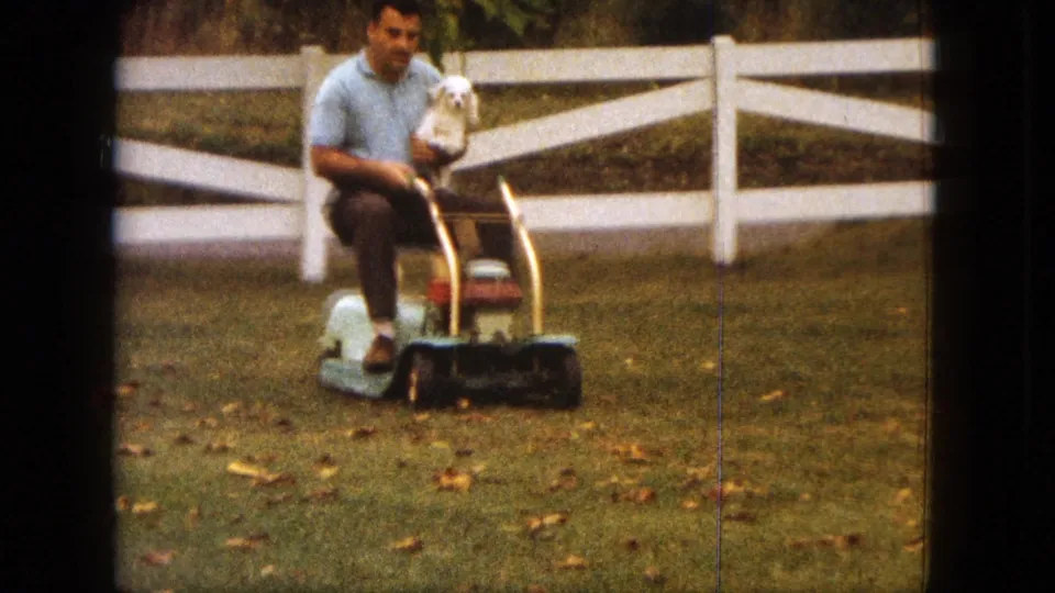 Front-facing 1963 Saint Paul frame shows the man holding the small white dog as the mower approaches across the grassy yard.