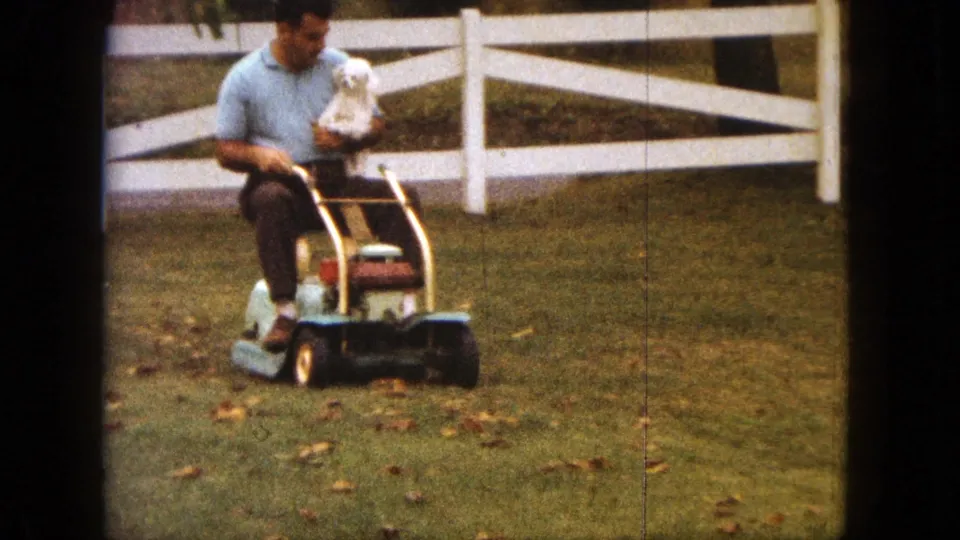 In Saint Paul, Minnesota in 1963, a man rides a small mower while holding a white pet dog beside a white fence.