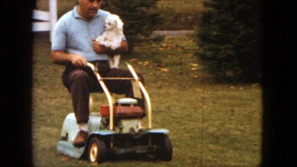 A closer 1963 home-movie view shows the man in a light blue polo holding the small white dog while riding the mower.