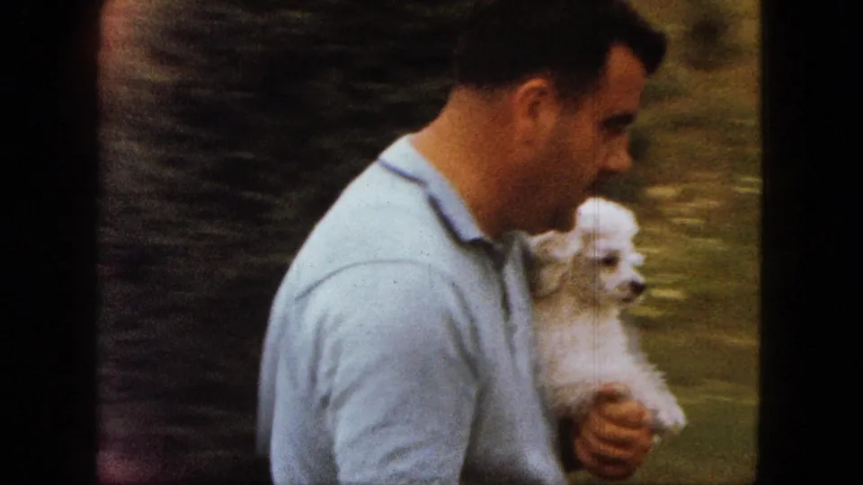 Close 1963 Saint Paul home-movie view of a man in a light blue polo holding a small white poodle against a green yard background.