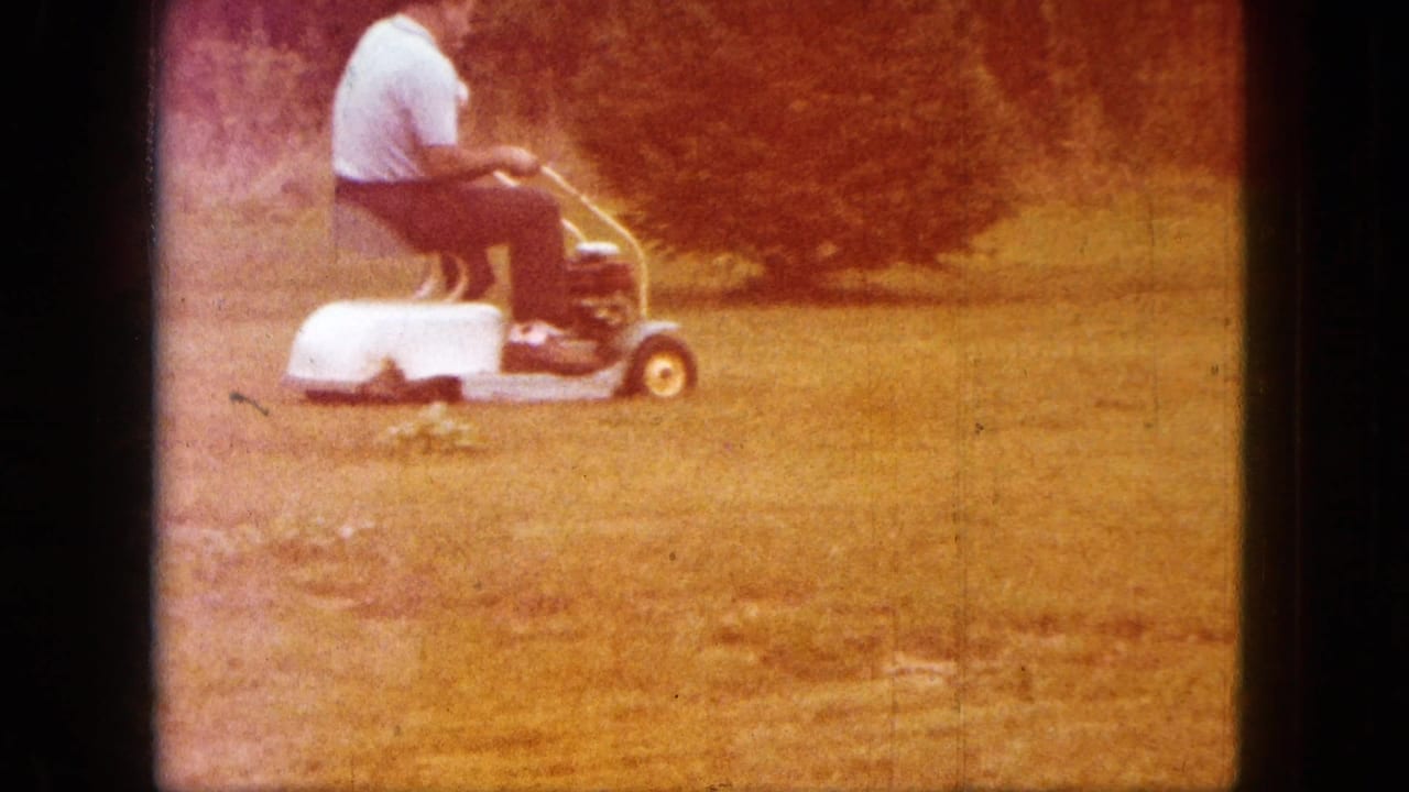 A man in a light shirt and dark pants rides a small mower across grass in Saint Paul, Minnesota, 1963 home-movie footage.