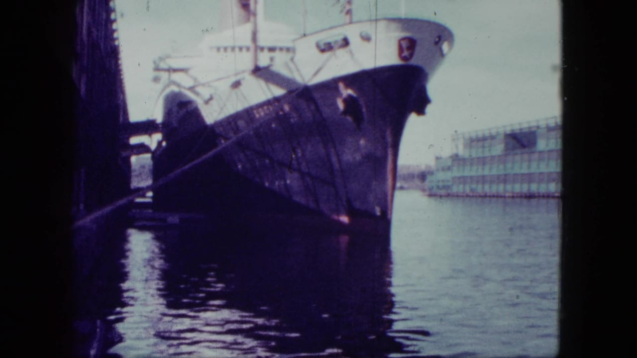 Large dark-hulled ship docked beside calm harbor water with a building in the background.