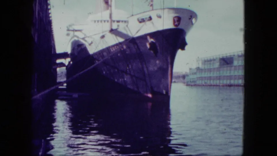 1963 San Diego home-movie frame showing the bow of a large docked ship, mooring lines, water reflections, and a waterfront building.