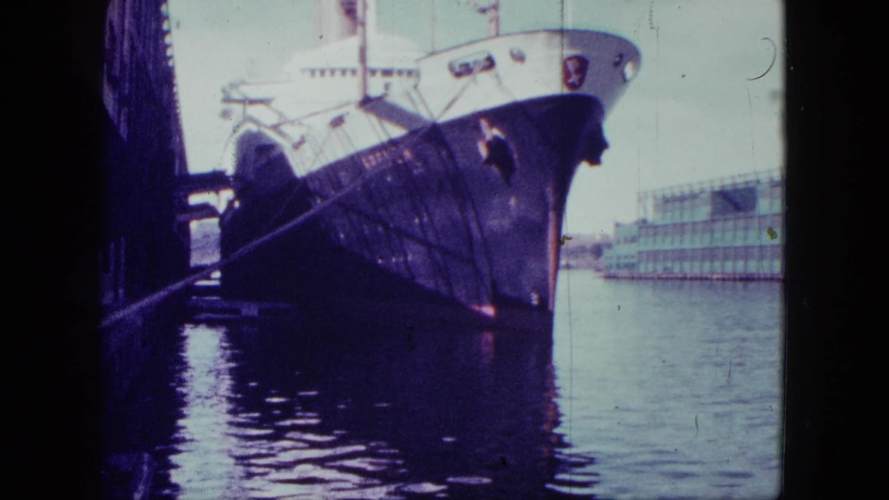 Bow of a large docked ship seen over calm water with a long building across the harbor.