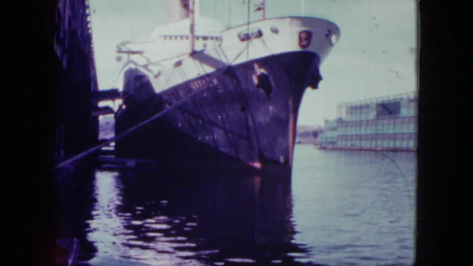 1963 San Diego 8mm frame showing a docked naval-looking vessel from close range, with purple color cast, harbor water, and background architecture.
