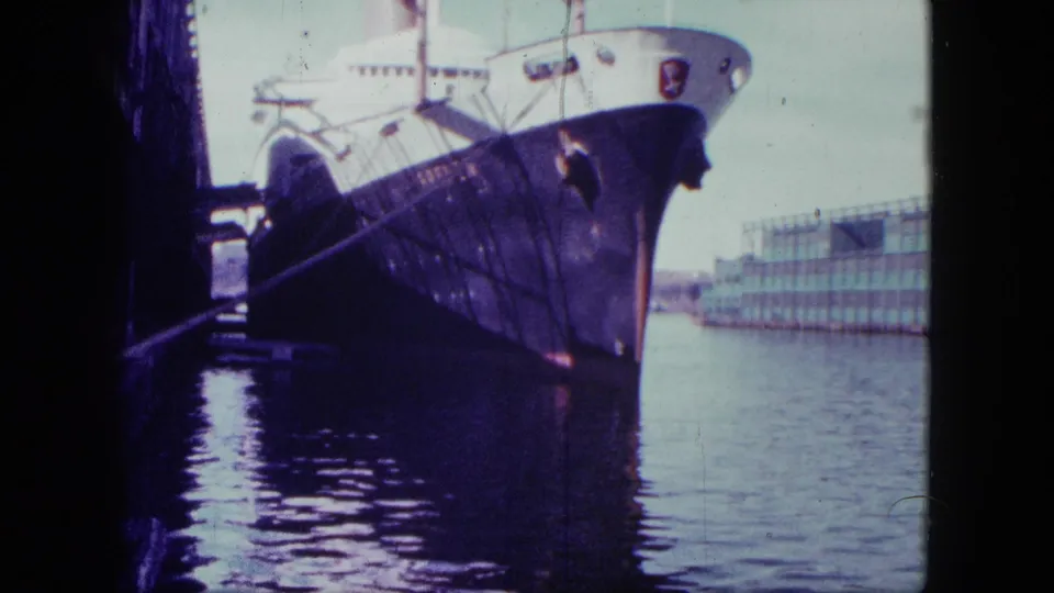 1963 San Diego home-movie frame showing a large ship moored at port, seen from a shaded dockside angle across harbor water.