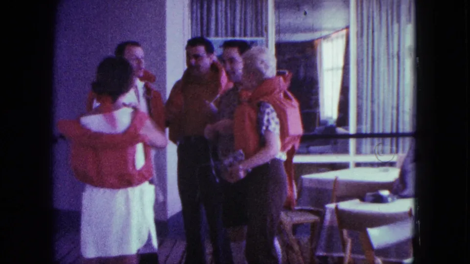 Ship passengers appear to chat informally during a life-jacket moment in a bright interior with tables, chairs, and curtained windows.