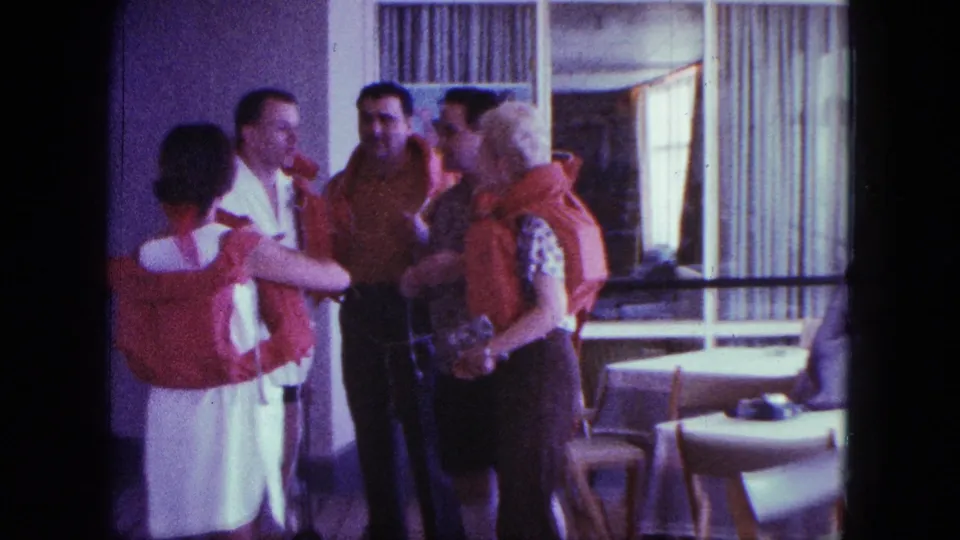 A final 1963 home-movie frame shows adults in orange life jackets gathered in a shipboard interior, continuing an informal conversation.