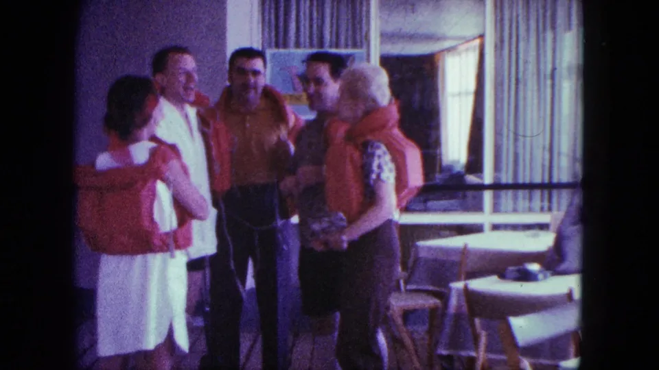 Adults in red life jackets converse in a bright San Diego interior, with tables, chairs, and curtains visible behind them.