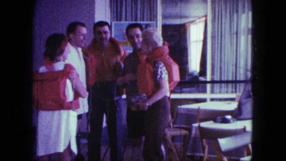 The 1963 home-movie frame shows five adults in life jackets talking closely in a sunlit dining area with tables and curtains.