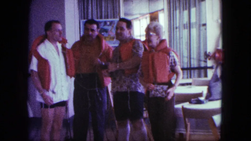 Adults in casual 1960s clothing stand in a shipboard room, several wearing orange-red life preservers near dining tables.