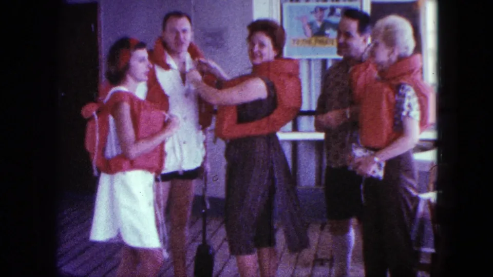 A woman assists a man with an orange life jacket during a 1963 San Diego boat-deck safety demonstration as others observe.
