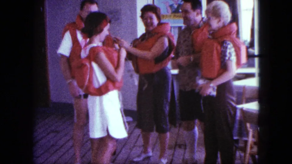 A 1963 San Diego boating group stands on a wooden deck as a woman continues demonstrating how a red life jacket fits.