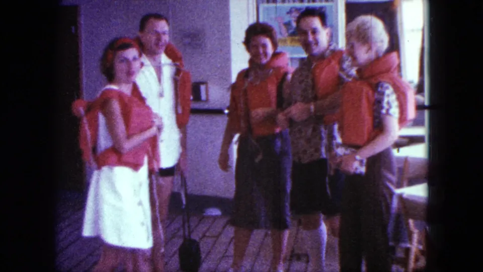 1963 San Diego home-movie frame with adults in red life jackets smiling or facing the camera on a deck after instruction.