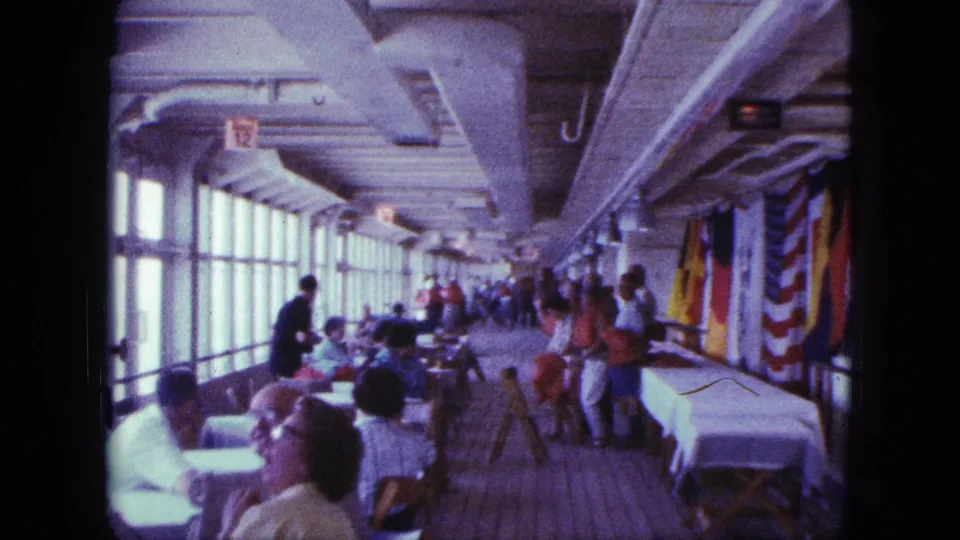 Boat interior shows passengers seated at tables beside long windows, with flags hanging along the opposite side.