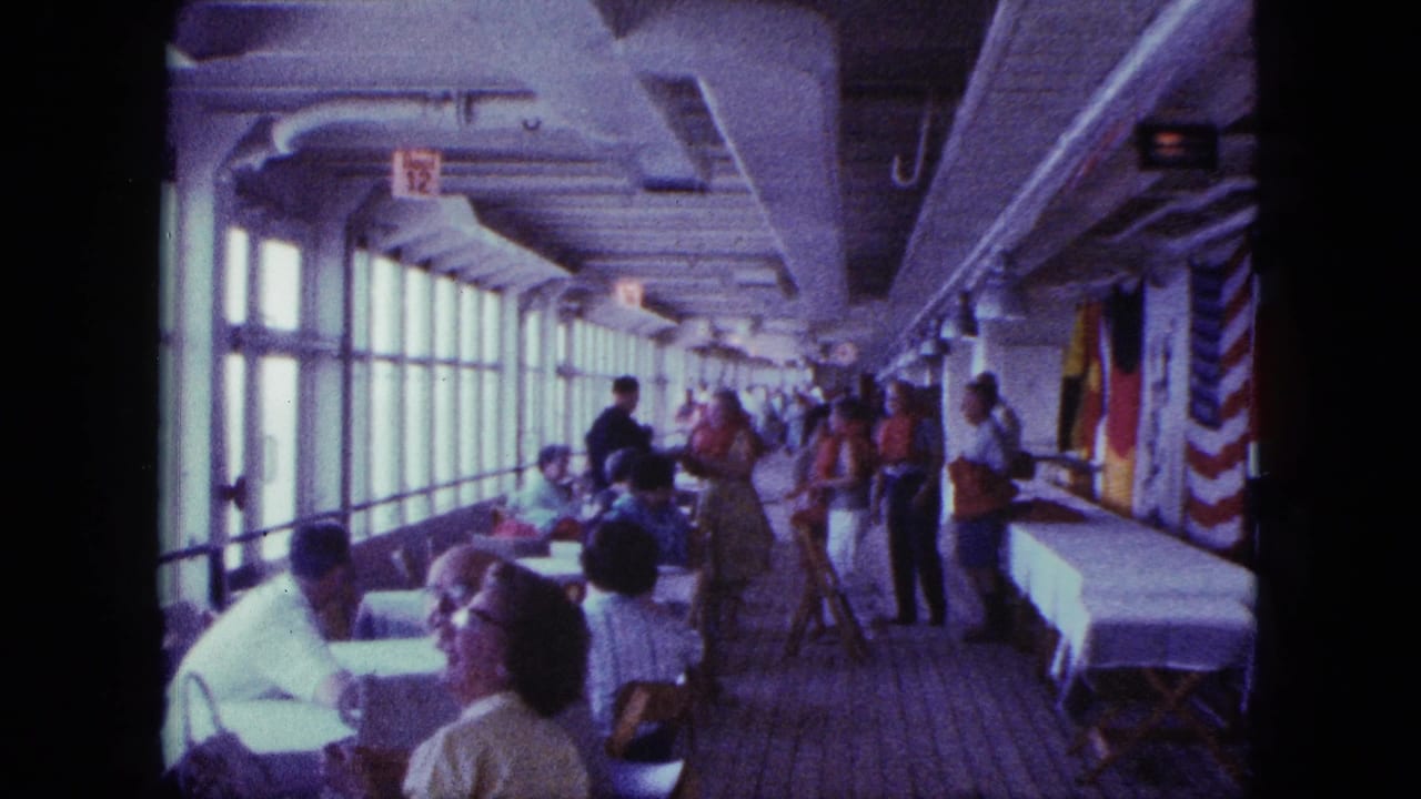 Passengers dine and stand along a window-lined ship deck with flags and a long table.