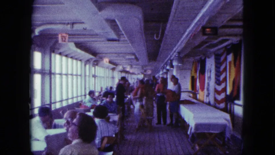 1963 San Diego home-movie view of passengers seated and standing along an enclosed ship deck with window-side tables and flag decorations.