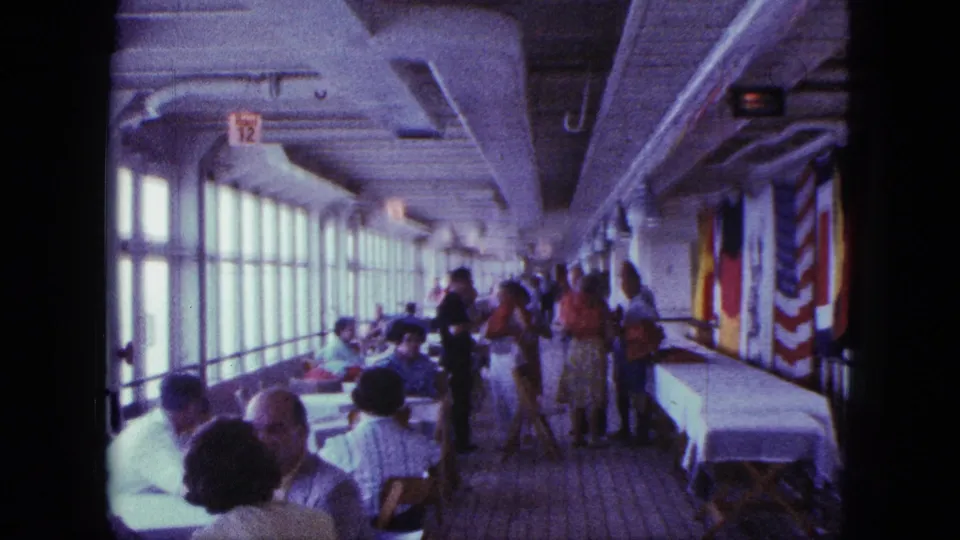 1963 San Diego shipboard scene with diners, standing passengers, flags, and a long covered table inside a bright enclosed deck.