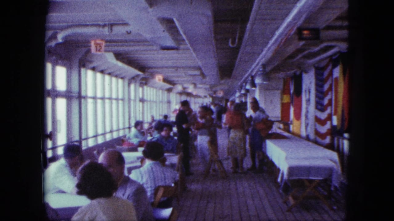 Passengers sit and stand in a long shipboard dining room with windows and hanging flags.