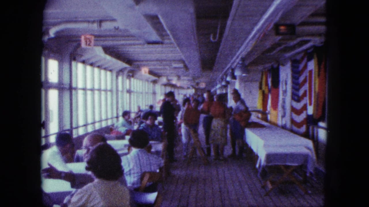 People dine and move through a shipboard room with windows, tables, and flags.